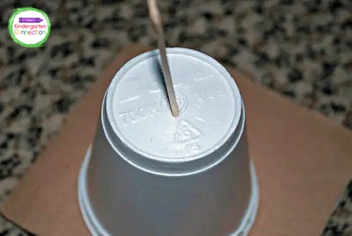 Cut a circular hole in the cardboard, glue the cup to one side, and slice a slit for the popsicle stick into the bottom of the cup.